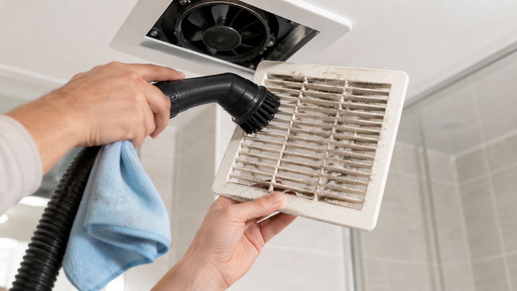 A person cleaning a dusty bathroom exhaust fan vent cover with a vacuum brush attachment
